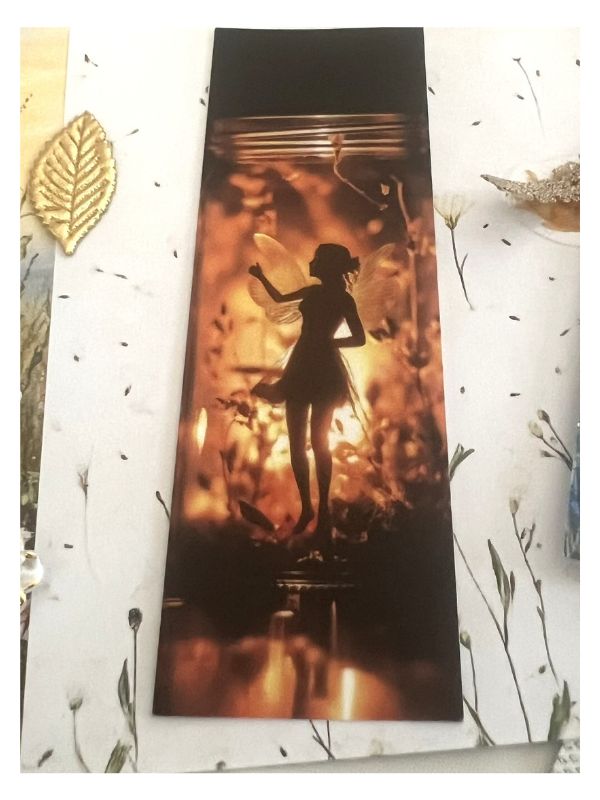 A rectangular printed card or bookmark featuring a dark silhouette of a fairy inside a glowing amber jar, placed on a white floral surface with decorative gold leaf accents.